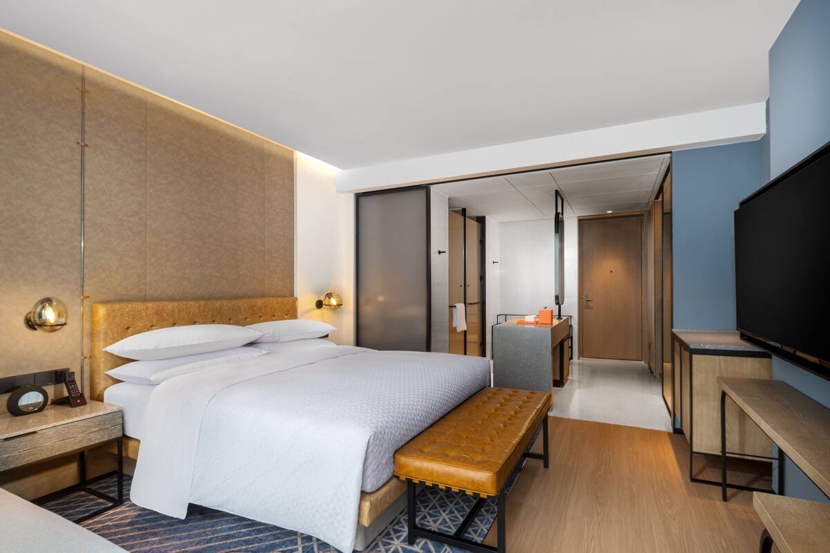 Ein modernes Hotelzimmer mit einem großen Bett, einem Nachttisch mit Lampe, einer Bank, einem Fernseher und einem Schreibtisch mit Computer.
