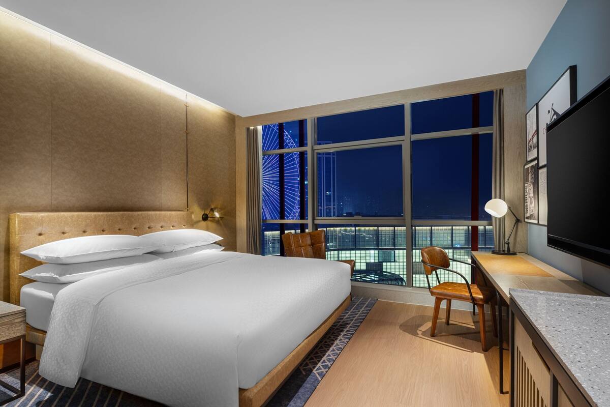 Ein modernes Hotelzimmer mit einem großen Bett, einem Schreibtisch und Blick auf eine beleuchtete Stadt bei Nacht.