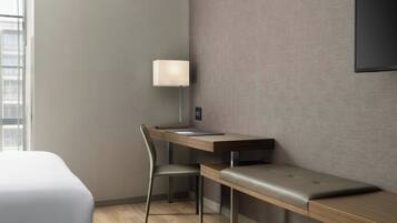 A modern hotel room with a wooden desk, a cushioned bench, a single chair, and a wall-mounted TV.
