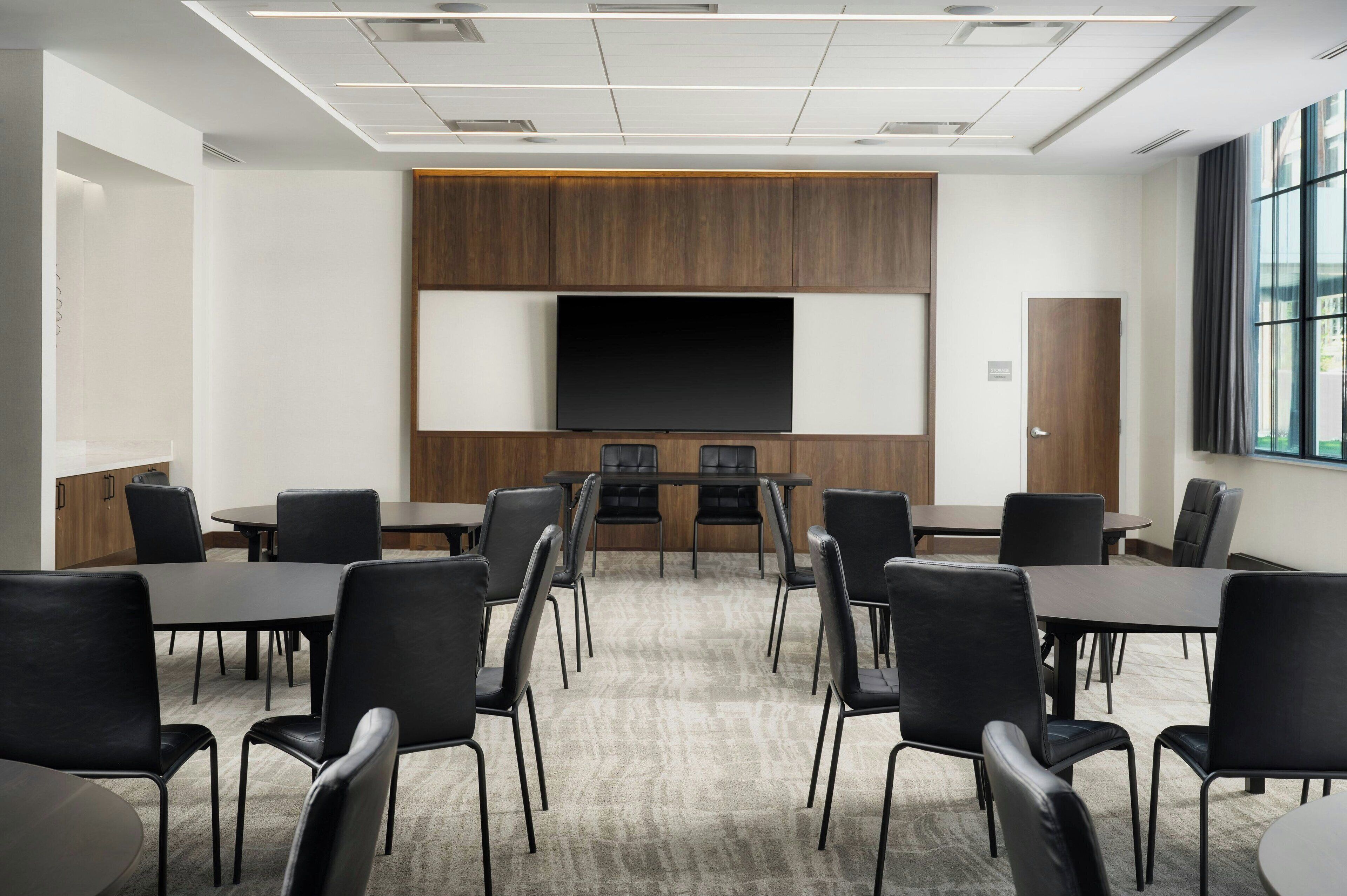 Un salón de conferencias con una gran pantalla plana, varias mesas y sillas de cuero negro.