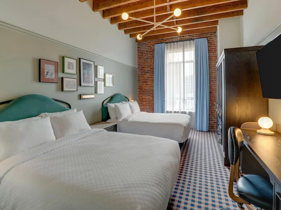 Habitación de hotel con dos camas, un escritorio con lámpara, pared de ladrillo y cuadros en la pared.