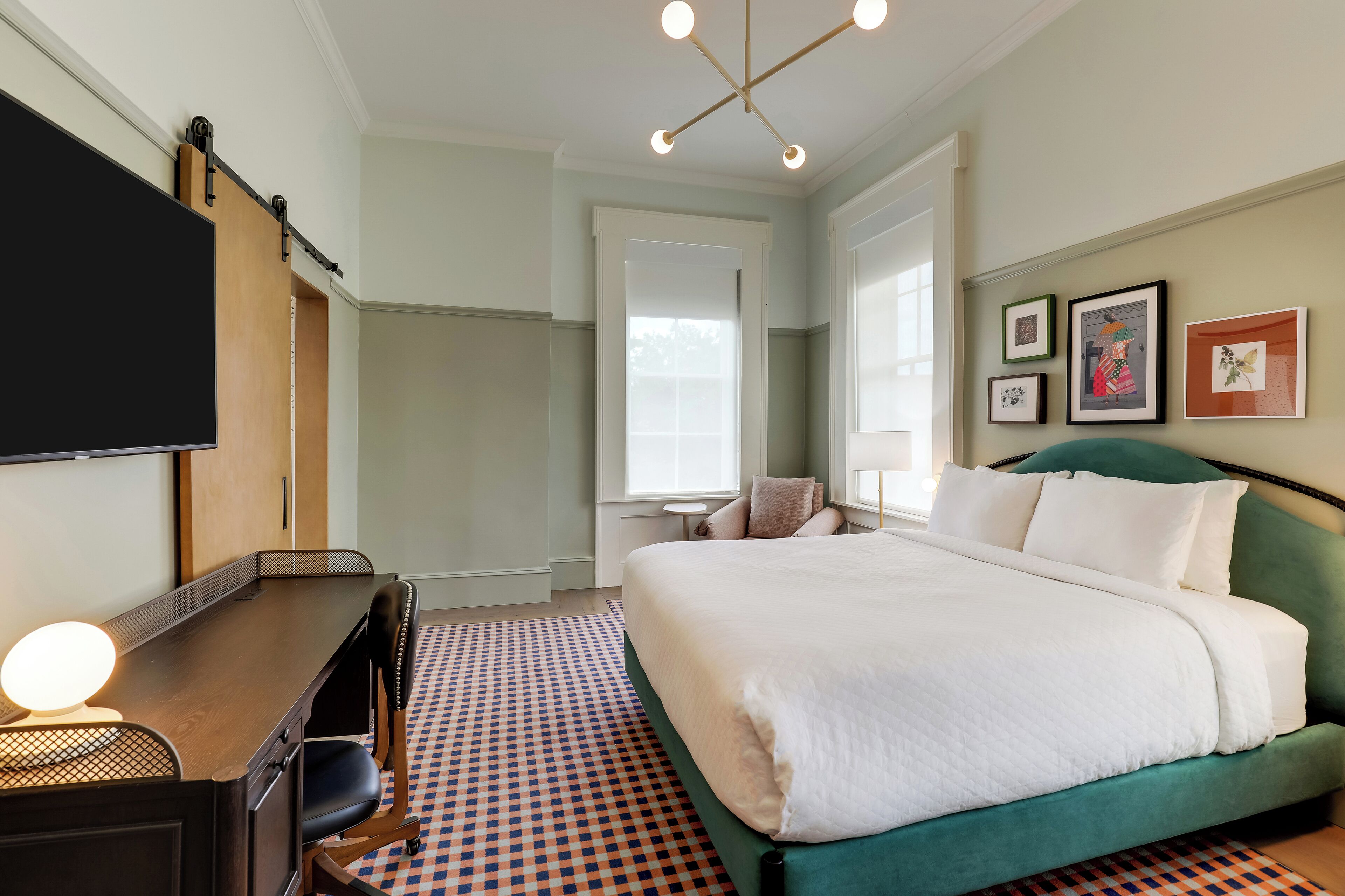 A hotel room with a bed, a desk, a chair, a TV, and framed pictures on the wall.