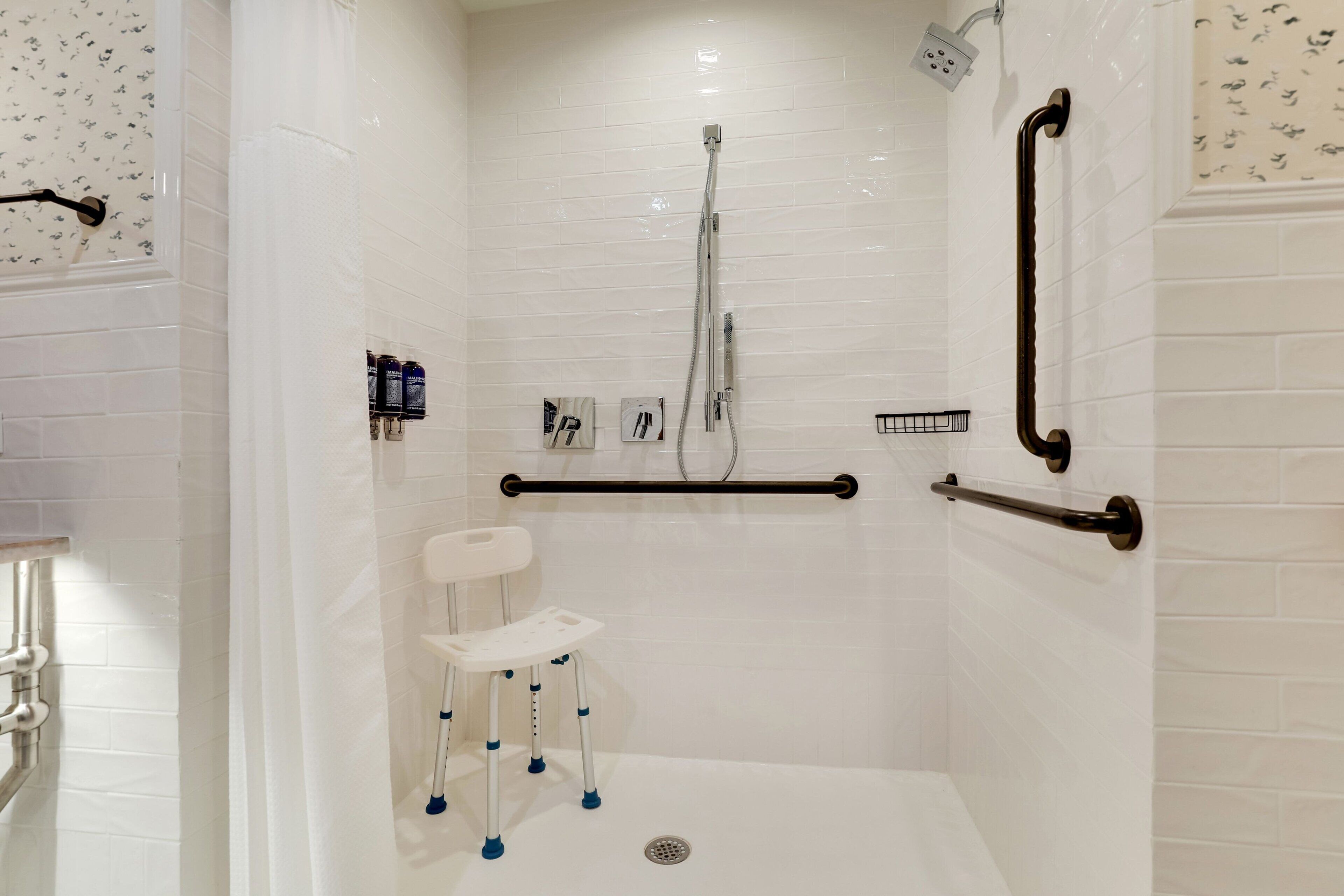 A walk-in shower with white tiled walls, a white shower seat, grab bars, and a handheld showerhead.