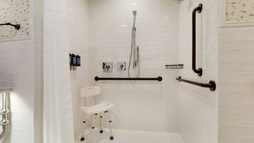 Ducha a nivel con paredes de azulejos blancos, asiento de ducha blanco, barras de apoyo y cabezal de ducha manual.