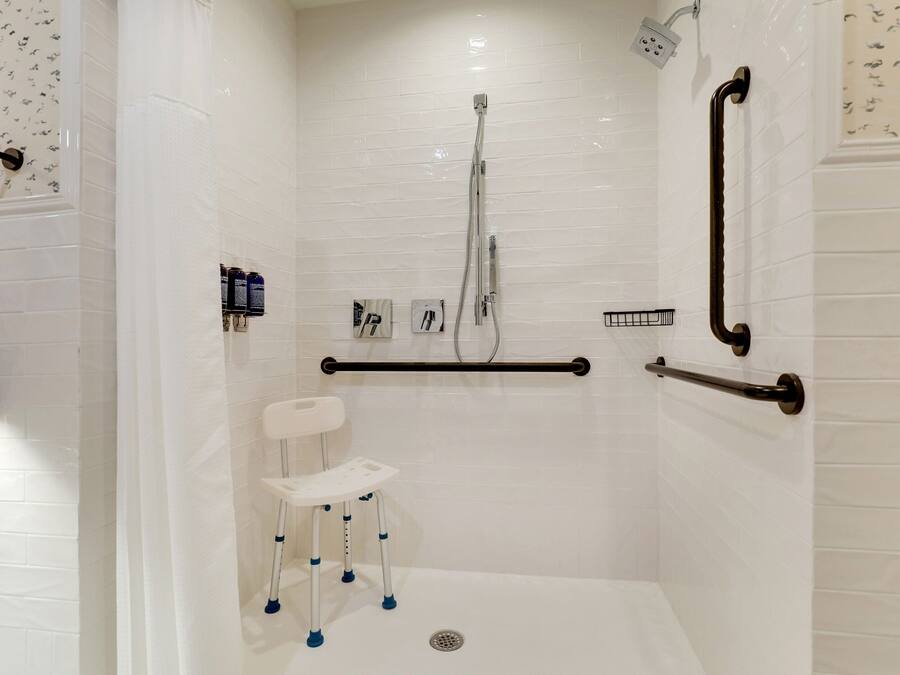 Ducha a nivel con paredes de azulejos blancos, asiento de ducha blanco, barras de apoyo y cabezal de ducha manual.