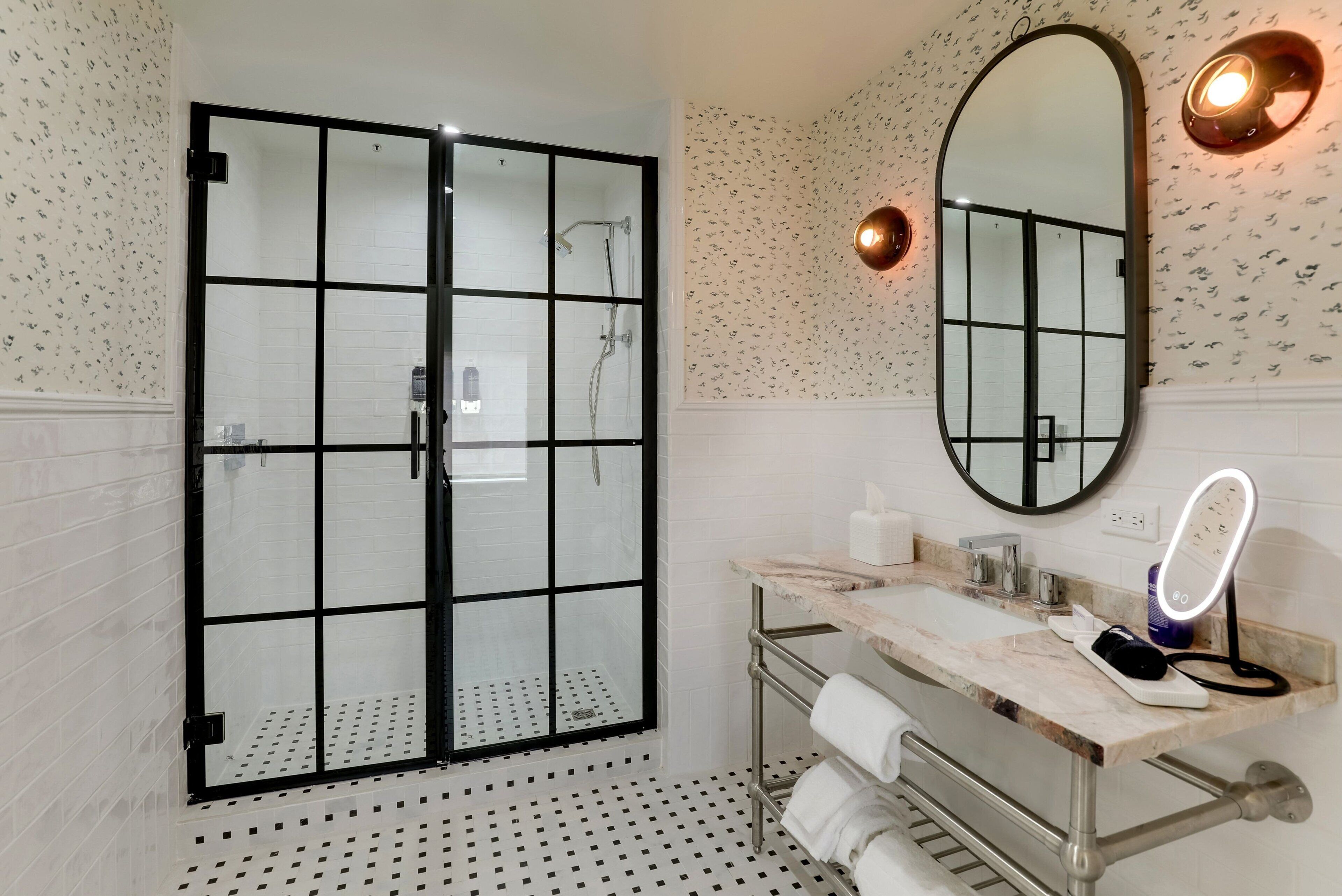 Een badkamer met een douchecabine van glas, een marmeren wastafel, een spiegel met verlichting en behang met een patroon.