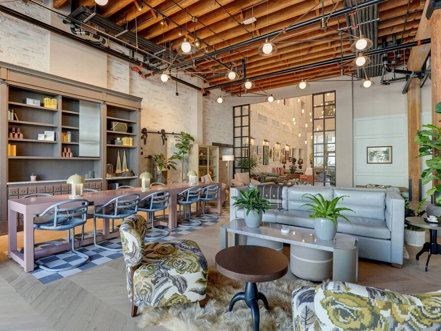 Un loft espacioso con vigas de madera a la vista, una zona de comedor con una mesa larga y sillas, un área de estar con sofás y una mesa de centro, y una amplia estantería llena de libros y objetos decorativos.