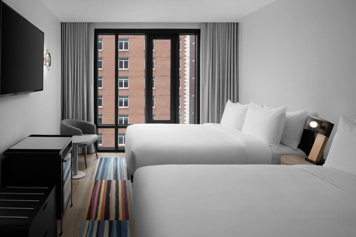 A hotel room with a bed, a desk, a chair, a TV, and a window with a view of buildings.
