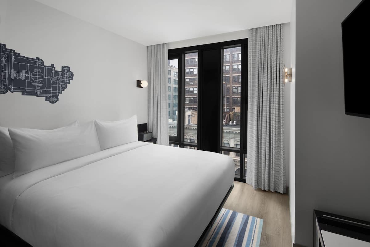 A modern hotel room with a large bed, a cityscape view through a window, and a wall-mounted artwork.