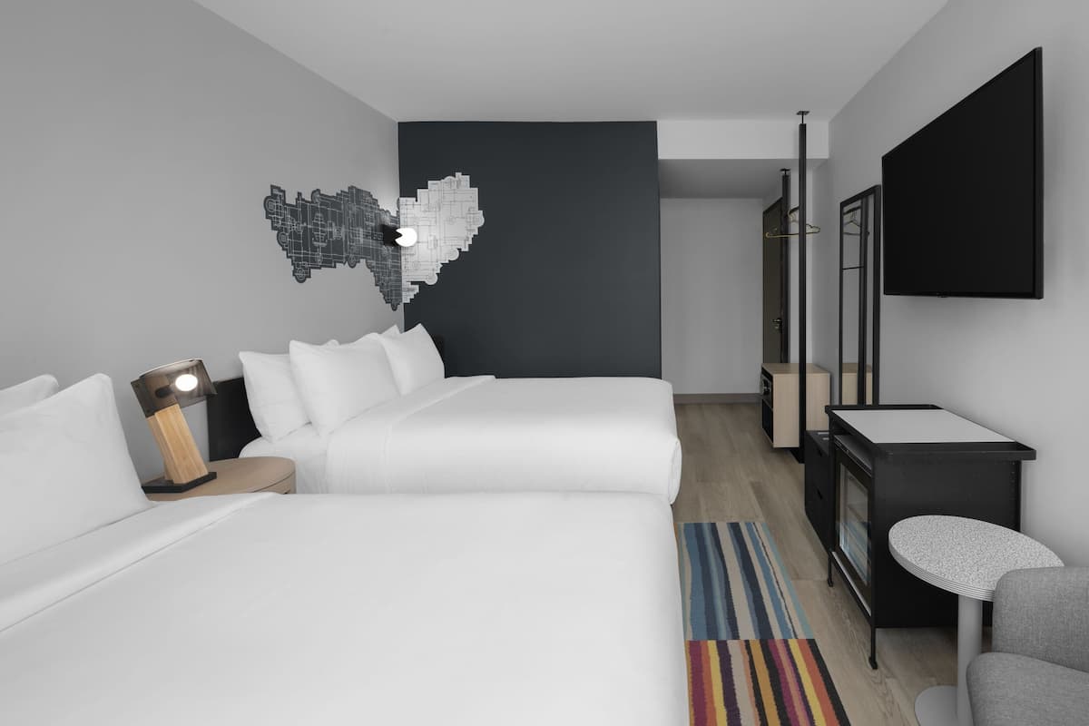 A modern hotel room with two beds, a desk, a mirror, and a wall art piece.
