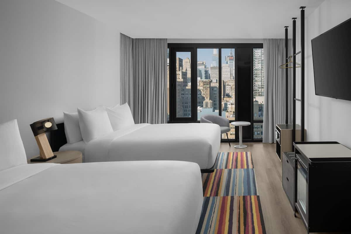 A hotel room with two beds, a desk, a chair, a TV, and a view of the cityscape.