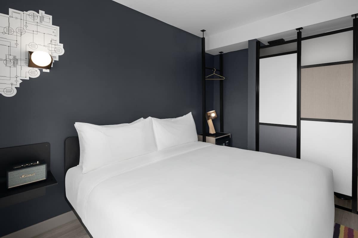 A modern hotel room with a large bed, a bedside table with a lamp, a wall-mounted light fixture, and a hanging garment rack.