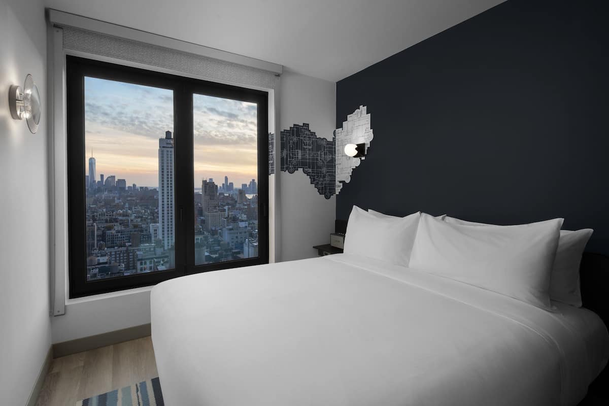 A modern hotel room with a large bed, a cityscape view through a window, and a unique wall art piece.