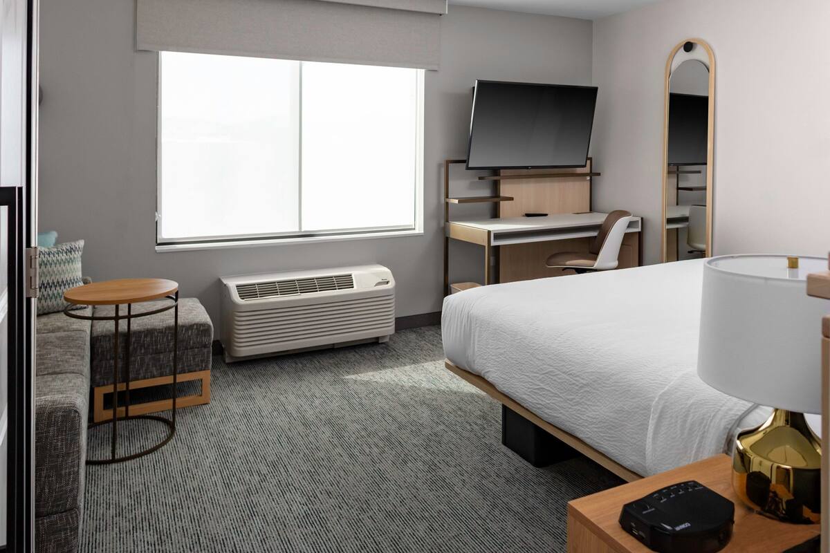 A hotel room with a bed, a desk with a chair, a TV, a mirror, a small round table, and a heater.