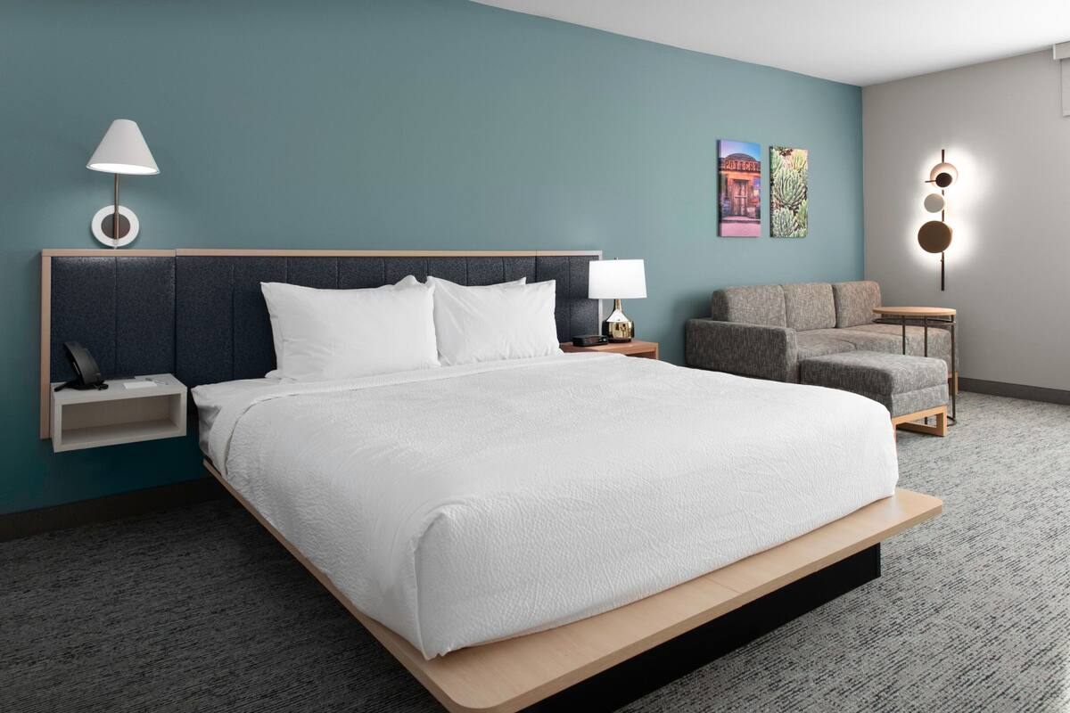 A modern hotel room with a large bed, a nightstand, a lamp, a sofa, and a small table.