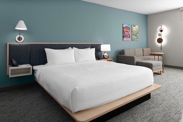 A modern hotel room with a large bed, a nightstand, a lamp, a sofa, and a small table. - TownePlace by Marriott Suites Buckeye Verrado (Buckeye)