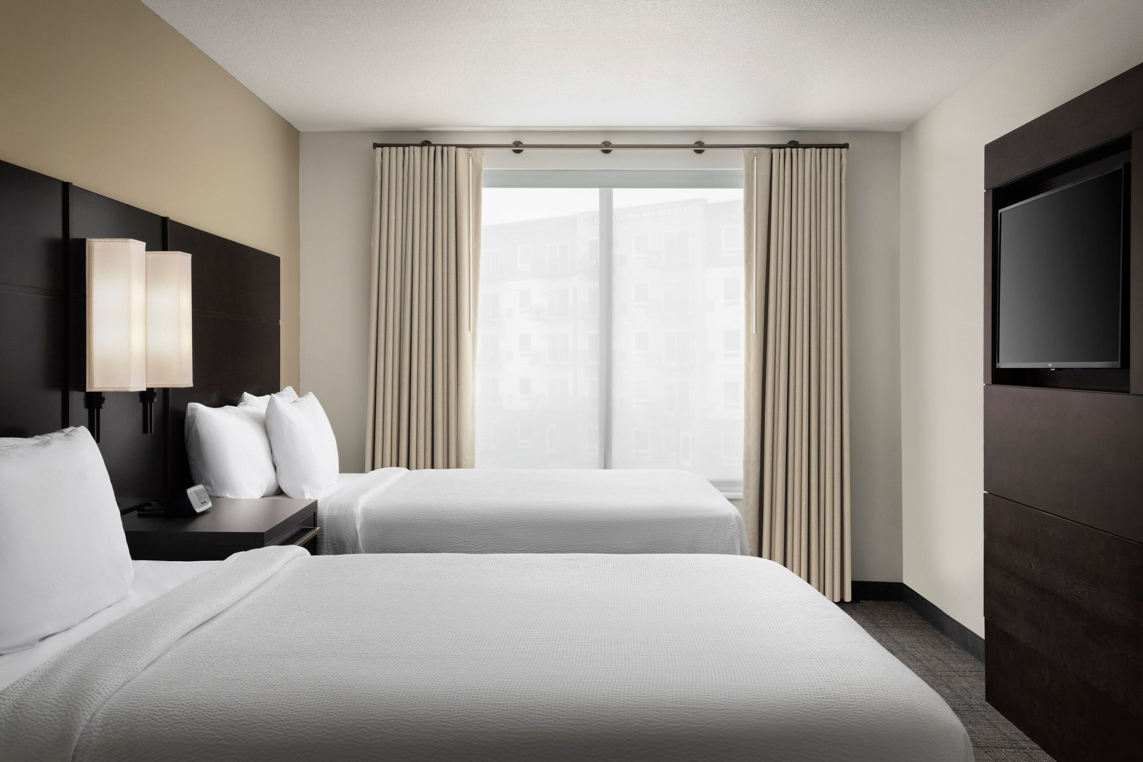 Ein Hotelzimmer mit zwei Betten, einem Fernseher, einem Nachttisch und einem Fenster mit Vorhängen.