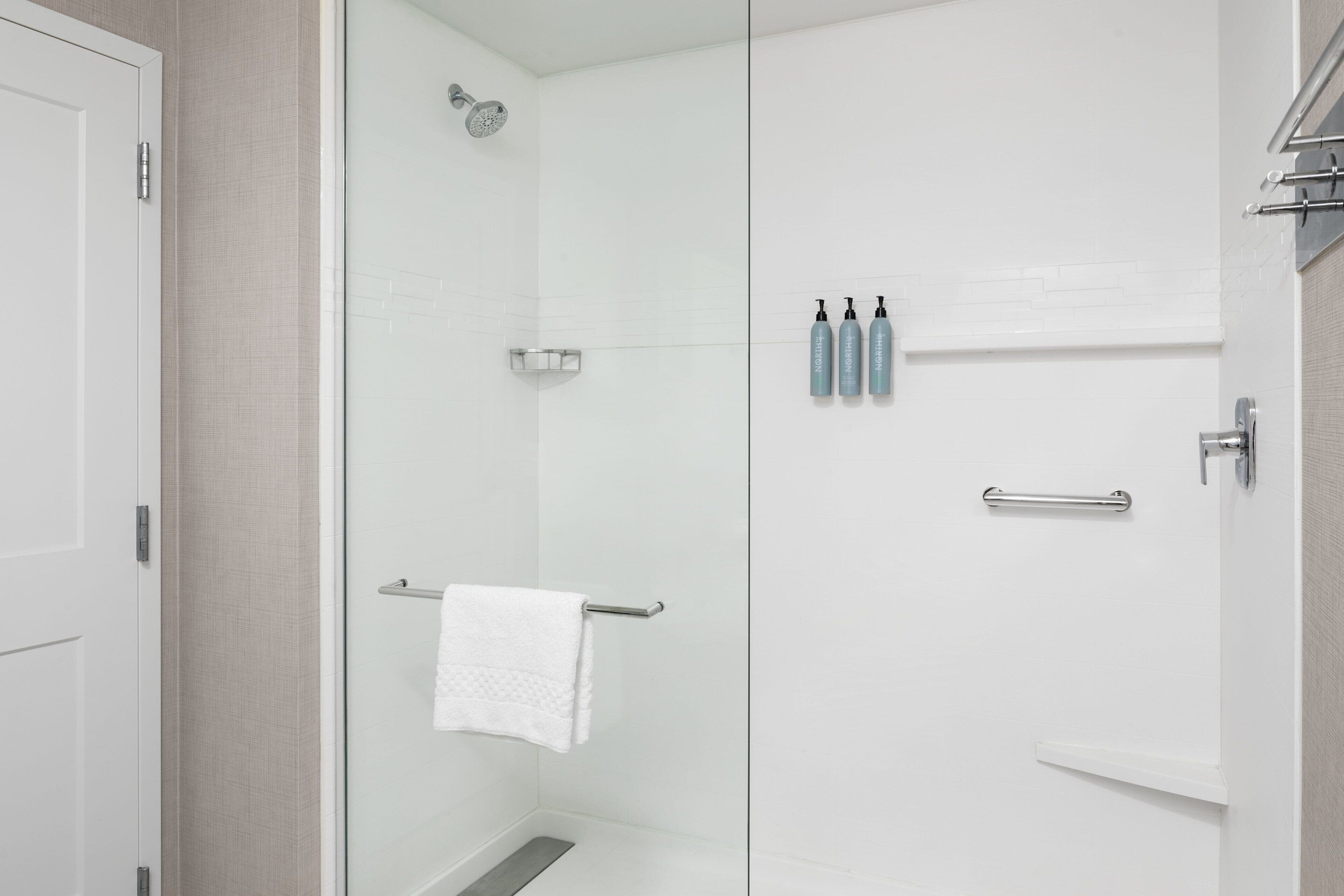 Une salle de bain moderne avec une douche à parois de verre, une serviette blanche suspendue à un porte-serviettes en métal et trois bouteilles de shampoing et d’après-shampoing posées sur le mur.