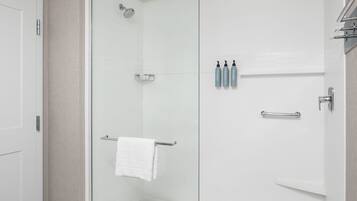Une salle de bain moderne avec une douche à parois de verre, une serviette blanche suspendue à un porte-serviettes en métal et trois bouteilles de shampoing et d’après-shampoing posées sur le mur.