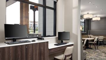Una habitación de hotel moderna con un escritorio, dos monitores, un teclado y un teléfono. Cuenta con un amplio ventanal que ofrece vistas de edificios. La habitación también tiene una zona de comedor con mesas y sillas.
