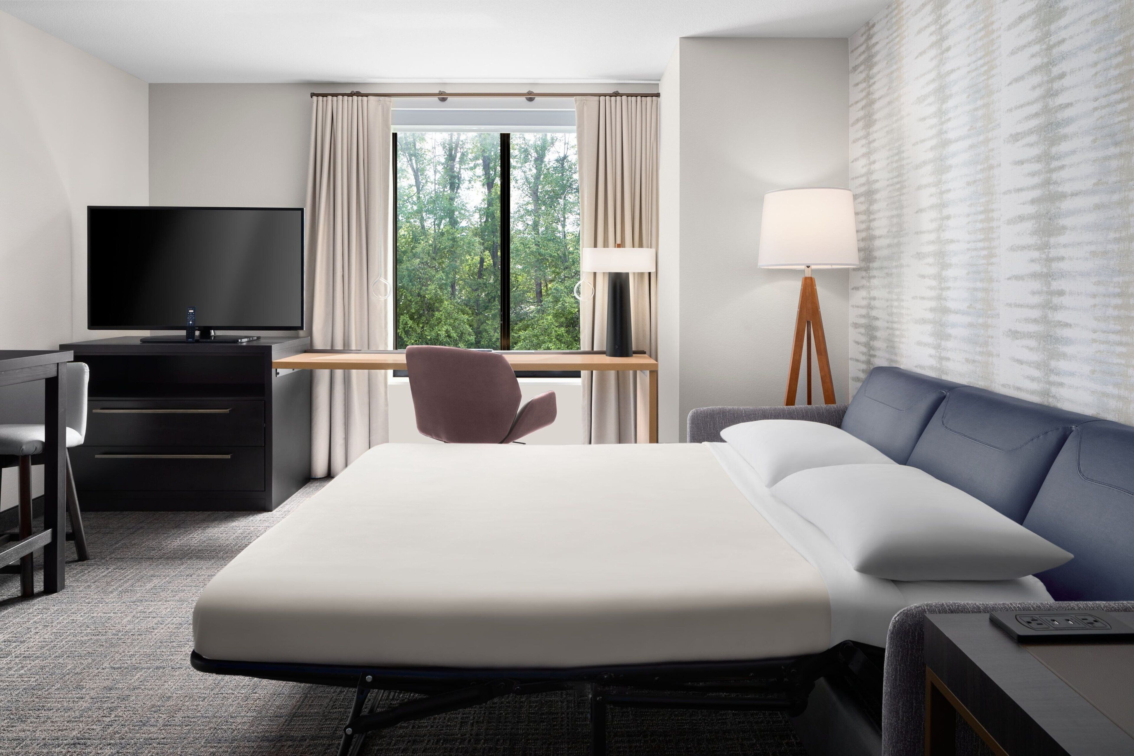 A hotel room with a bed, a desk, a chair, a TV, and a lamp.