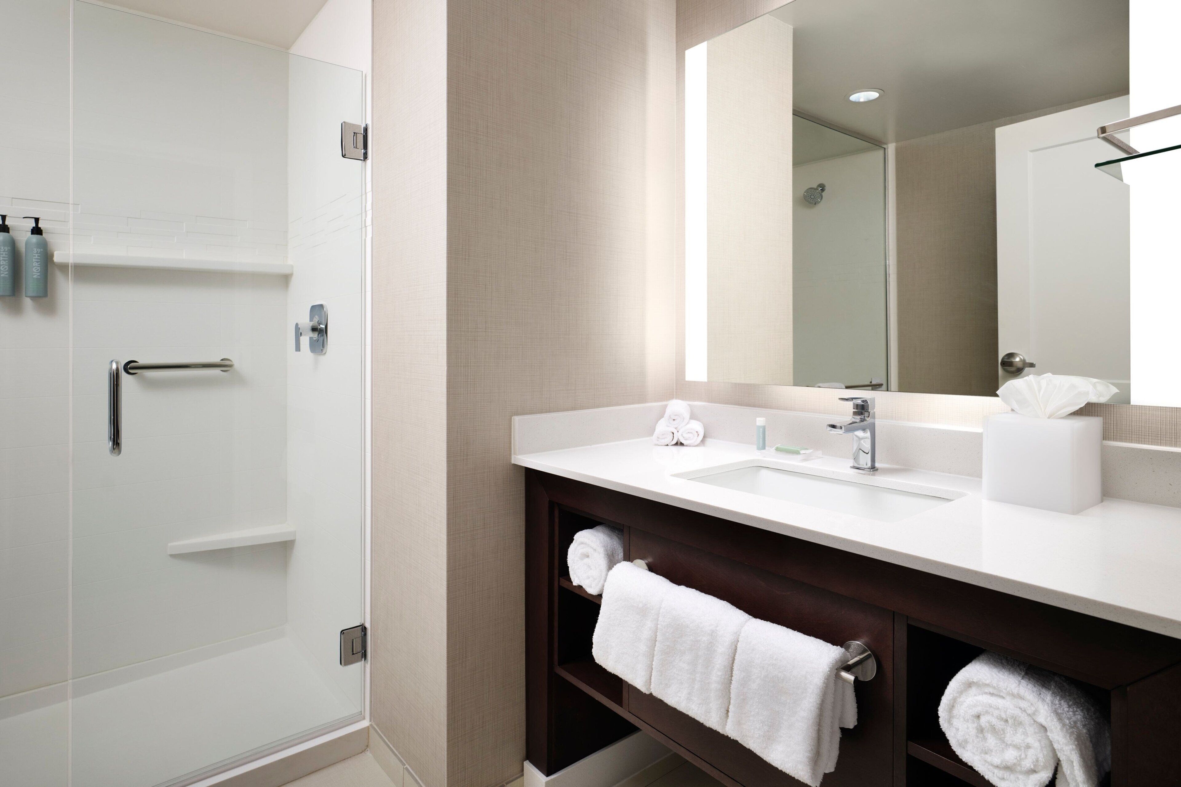 A modern bathroom with a walk-in shower, a white sink with a mirror above, and a dark wooden vanity with towels.