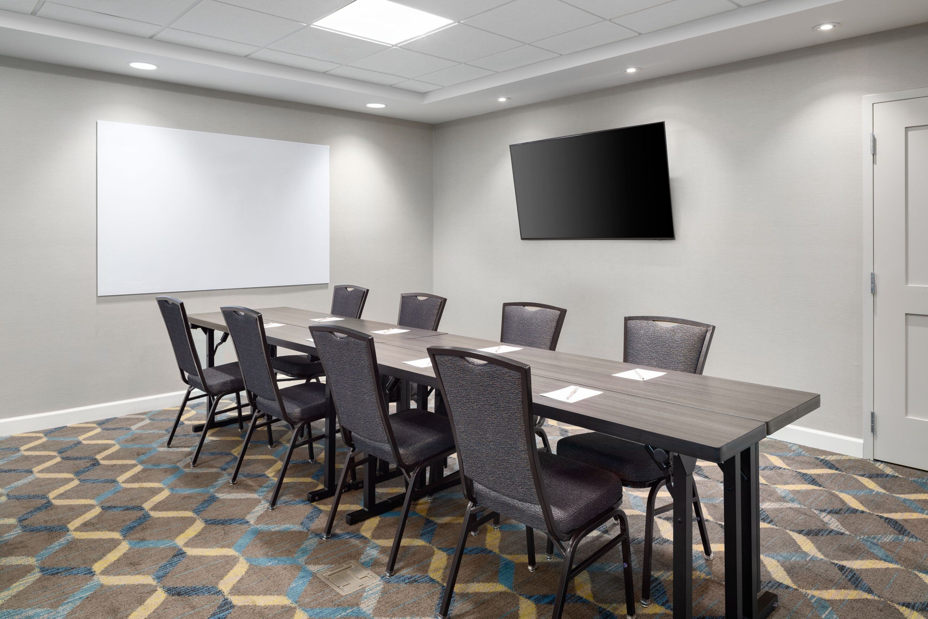 Un salón de conferencias con una larga mesa, seis sillas, una pizarra y un televisor de pantalla plana en la pared.