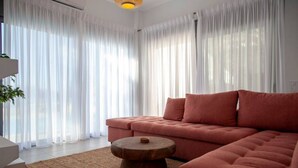 A modern living room with a sectional sofa, a round wooden coffee table, and sheer white curtains. - Villa Petalouda Paleros (Aktio-Vonitsa)