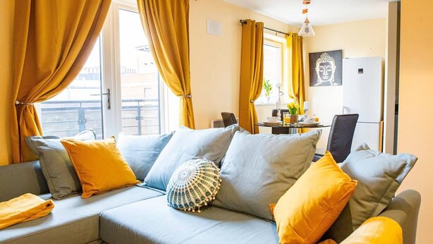 A modern living room with a grey sectional sofa, yellow cushions, and a dining area with a table set for two. - Inviting 2-bed Apartment in Wolverhampton (Wolverhampton)