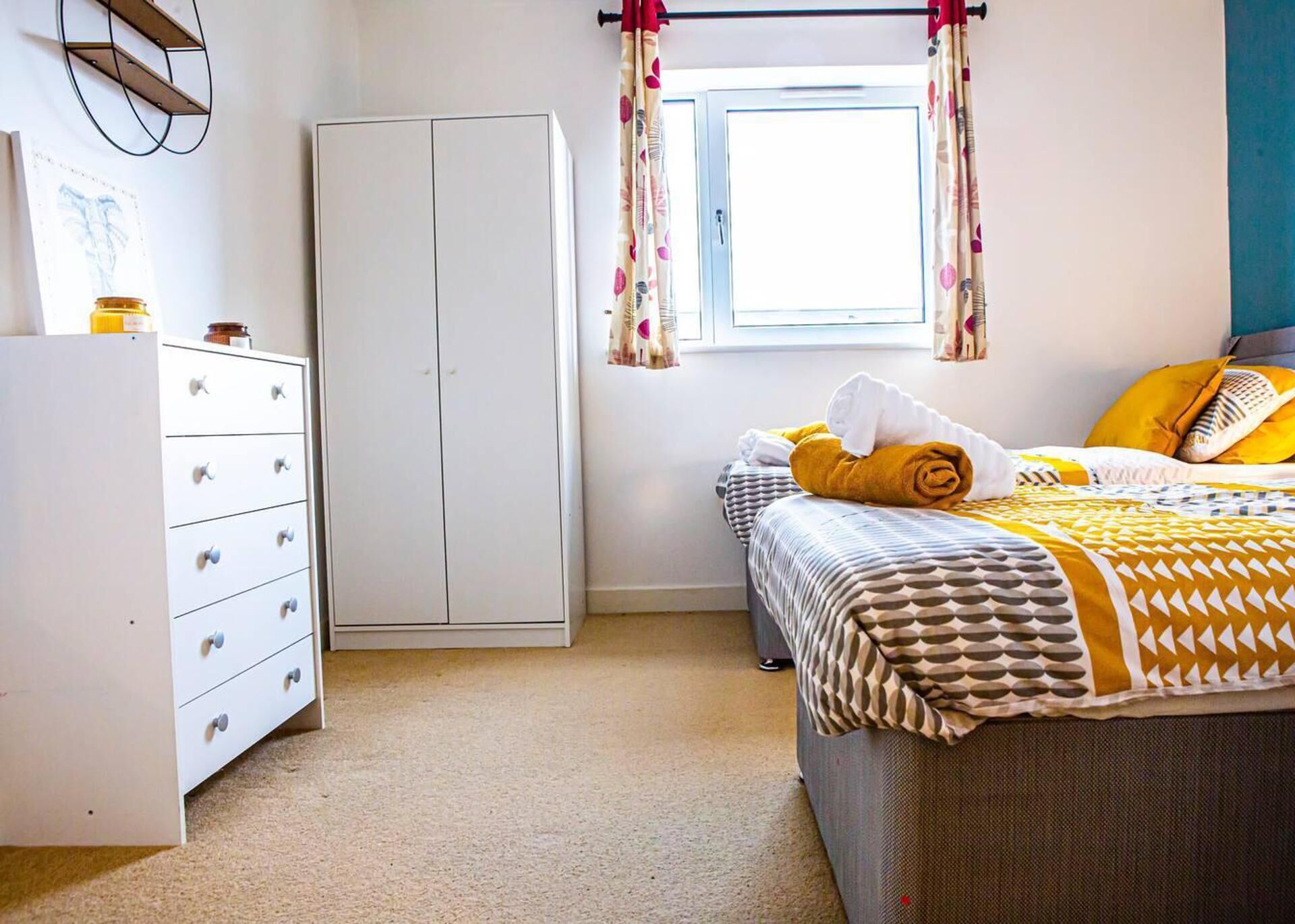 A bedroom with a bed, a wardrobe, a window with curtains, and a small shelf with items.