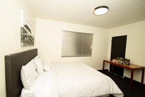 Ett hotellrum med en säng, ett sängbord, ett fönster med persienner, en tavla som hänger på väggen och en dörr.