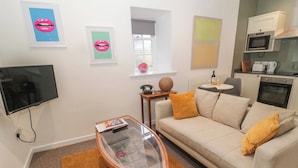 Una sala de estar con un sofá, una mesa de centro de vidrio, un televisor y una zona de cocina con microondas y horno.