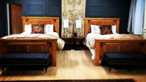 Um quarto com duas camas de madeira, um banco acolchoado azul escuro, uma pequena mesa com abajur e um tapete de parede com padrão floral.