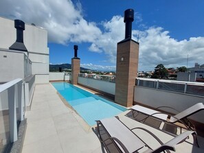 A rooftop pool with lounge chairs, a clear sky, and distant buildings. - Ap Madri - Piscina - Floripa - Conforto (Florianópolis)