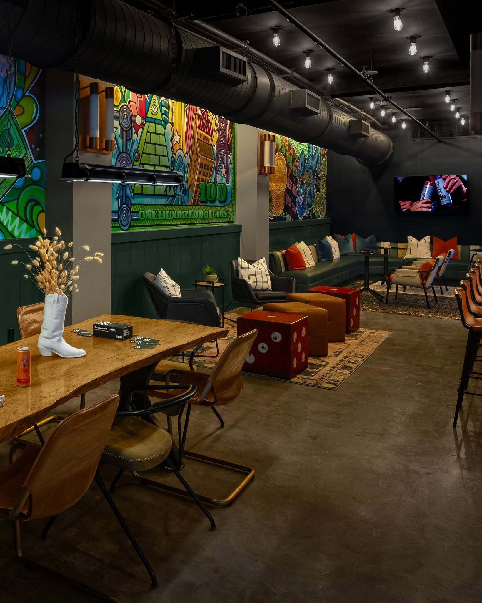Un'area lounge moderna con un lungo tavolo di legno, sedute imbottite e murales colorati alle pareti.