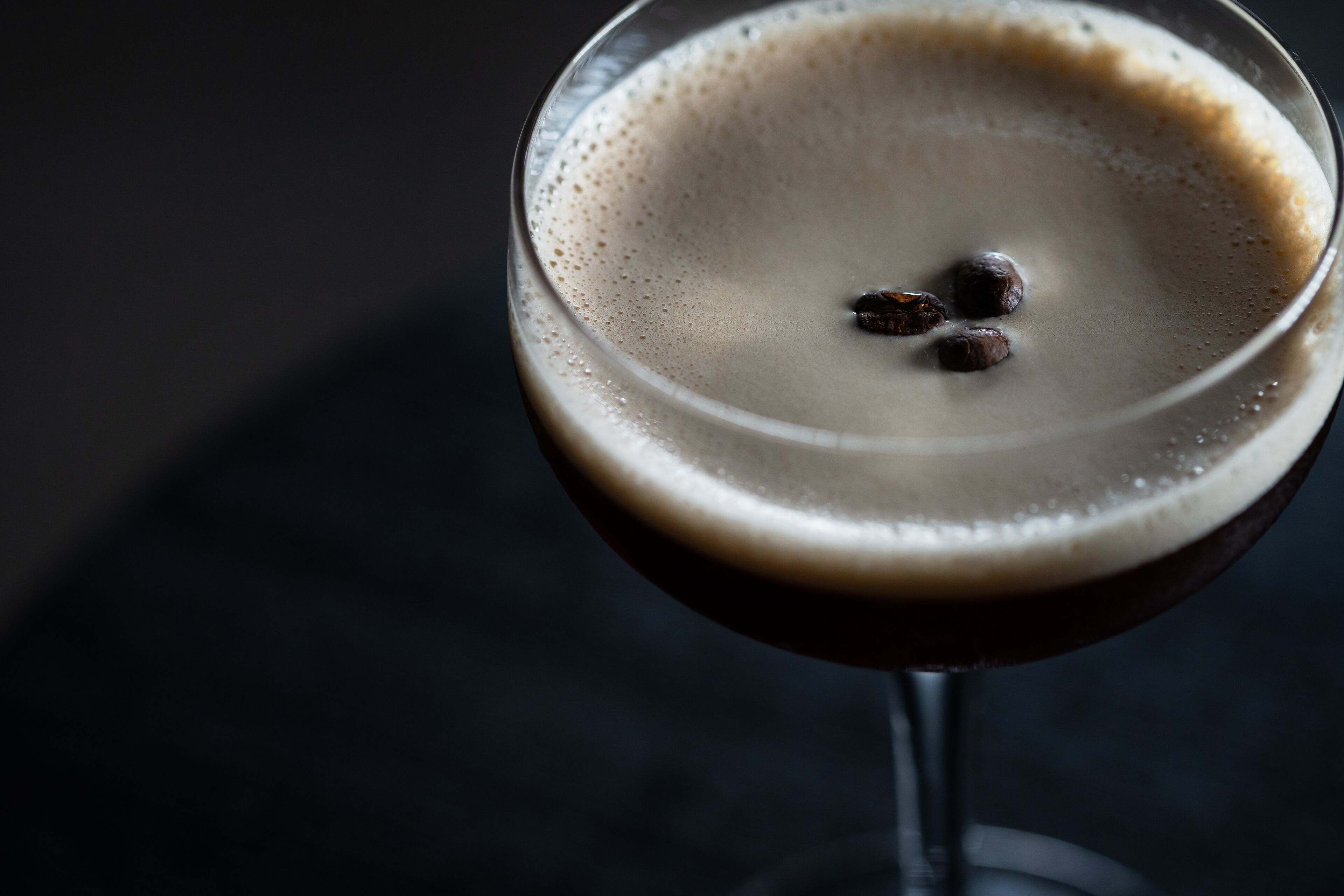 Een close-up van een donker drankje met schuim en enkele koffiebonen erbovenop, geserveerd in een helder glas.