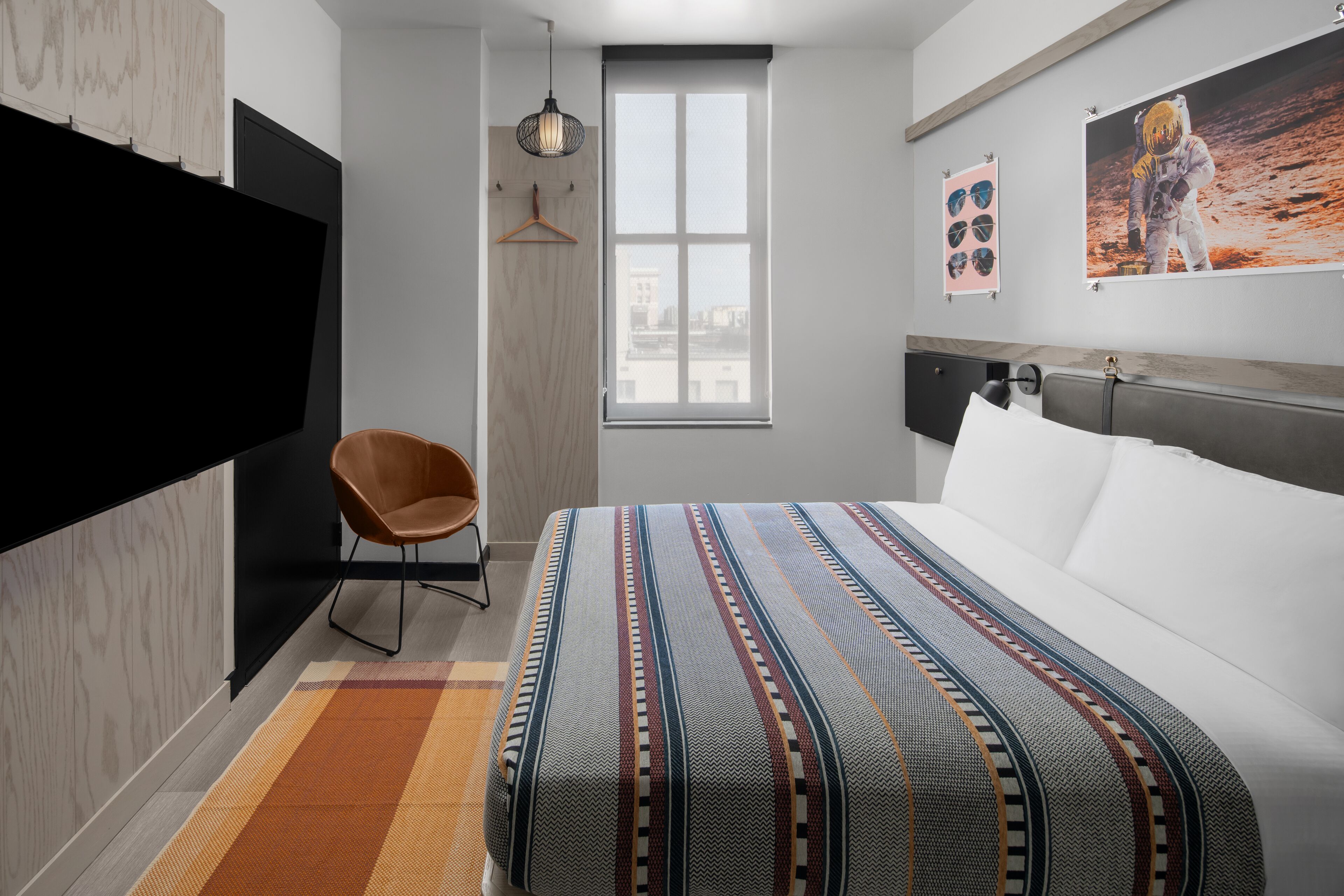 A modern bedroom with a large bed, a hanging light fixture, a chair, and a window with a city view.
