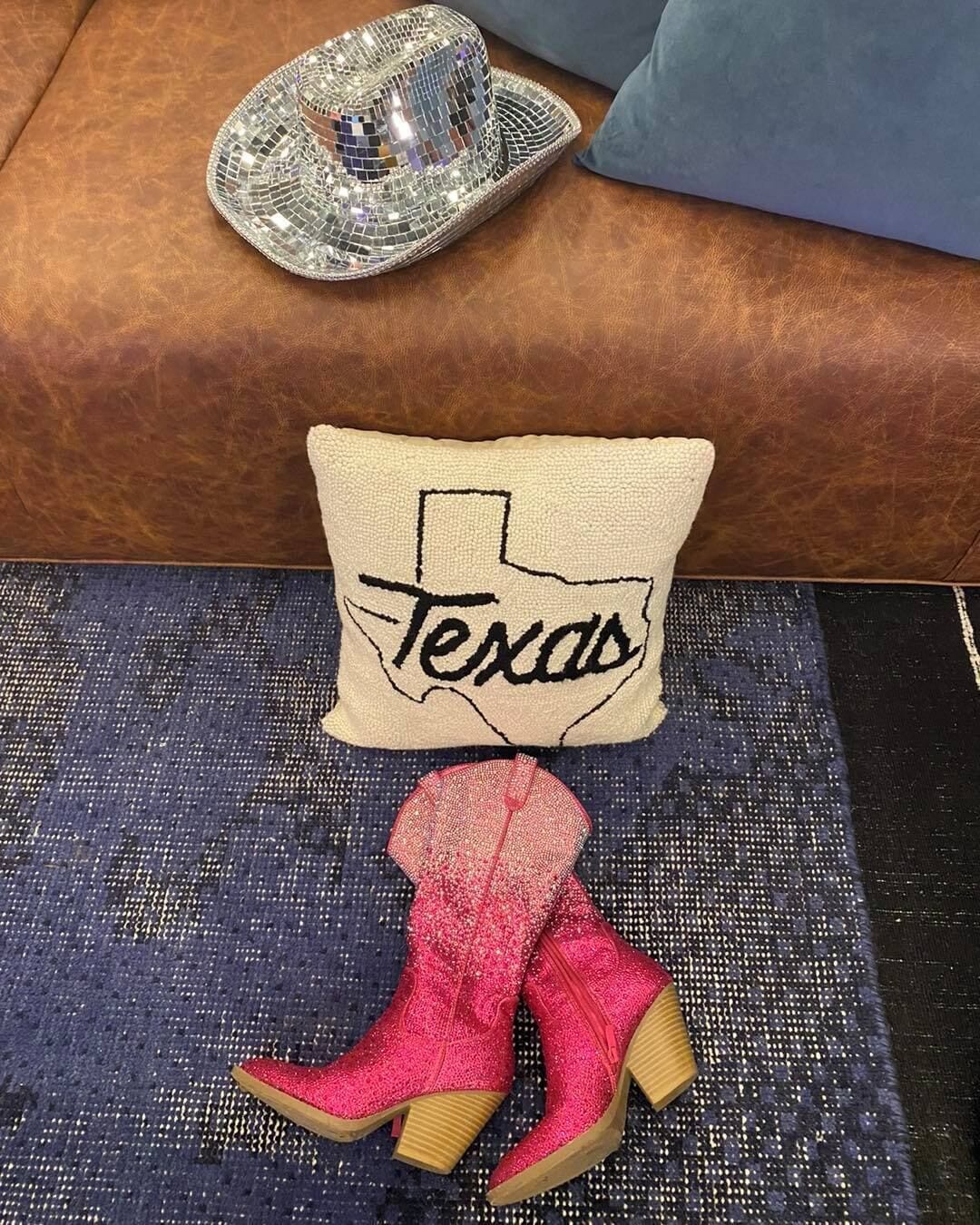 A decorative pillow with "Texas" on it, a pair of pink glittery boots, and a shiny cowboy hat on a textured surface.