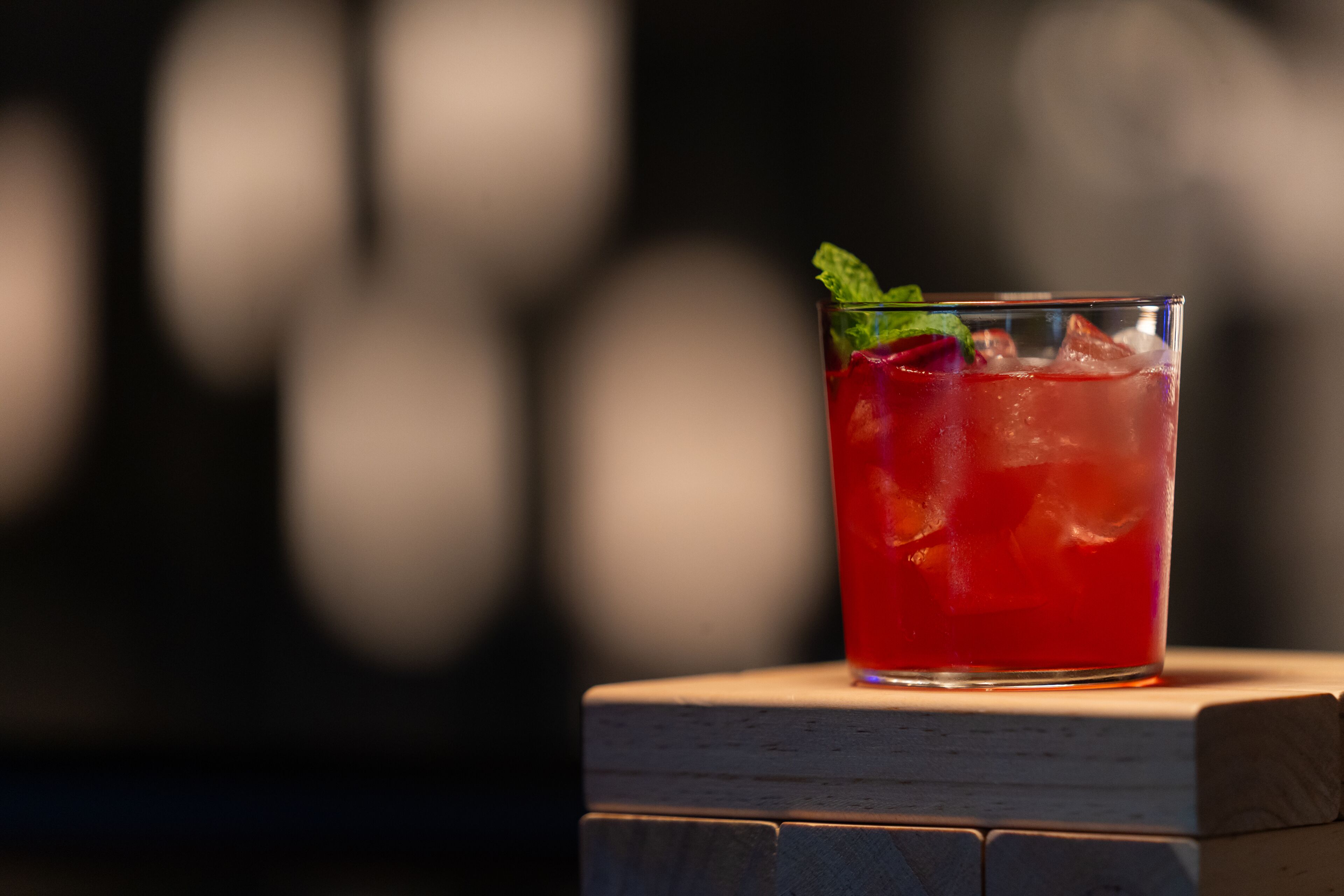 A glass of red drink with ice and a mint leaf on a wooden surface.