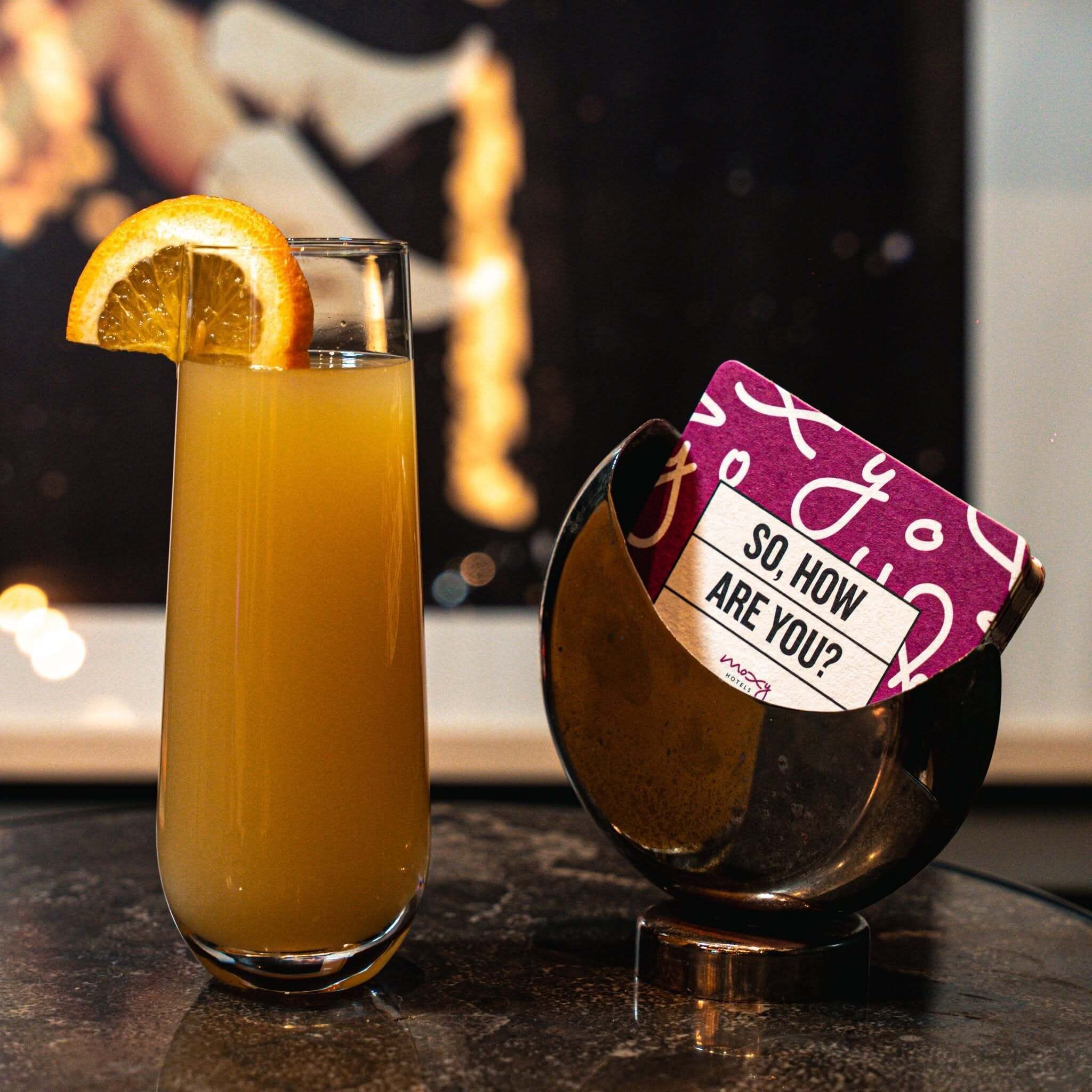 Un vaso alto con una bebida amarillenta y una rodaja de naranja como decoración, junto a un objeto metálico con una etiqueta.