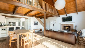 Een ruime woonkamer met houten plafond, een open haard, een eethoek met tafel en stoelen, en een keuken op de achtergrond.
