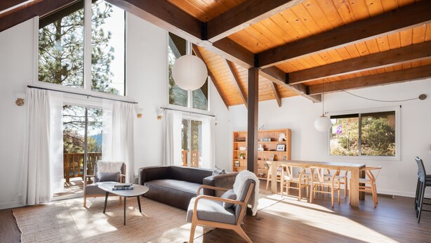 Een ruime woonkamer met houten plafond, grote ramen, een bank, een eettafel en een stoel.