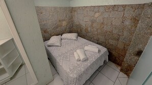 A small, narrow room with a bed against a textured stone wall, a white shelf unit, and tiled flooring. - Loft Barra da Fortaleza - Piscina (Florianópolis)