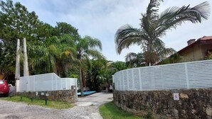 A gated entrance with a stone wall and a metal fence, leading to a pool area surrounded by palm trees and a clear sky. - Loft Barra da Fortaleza - Piscina (Florianópolis)