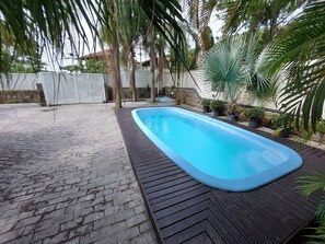 A rectangular swimming pool with a wooden deck surrounding it, set in a landscaped garden with palm trees and potted plants.