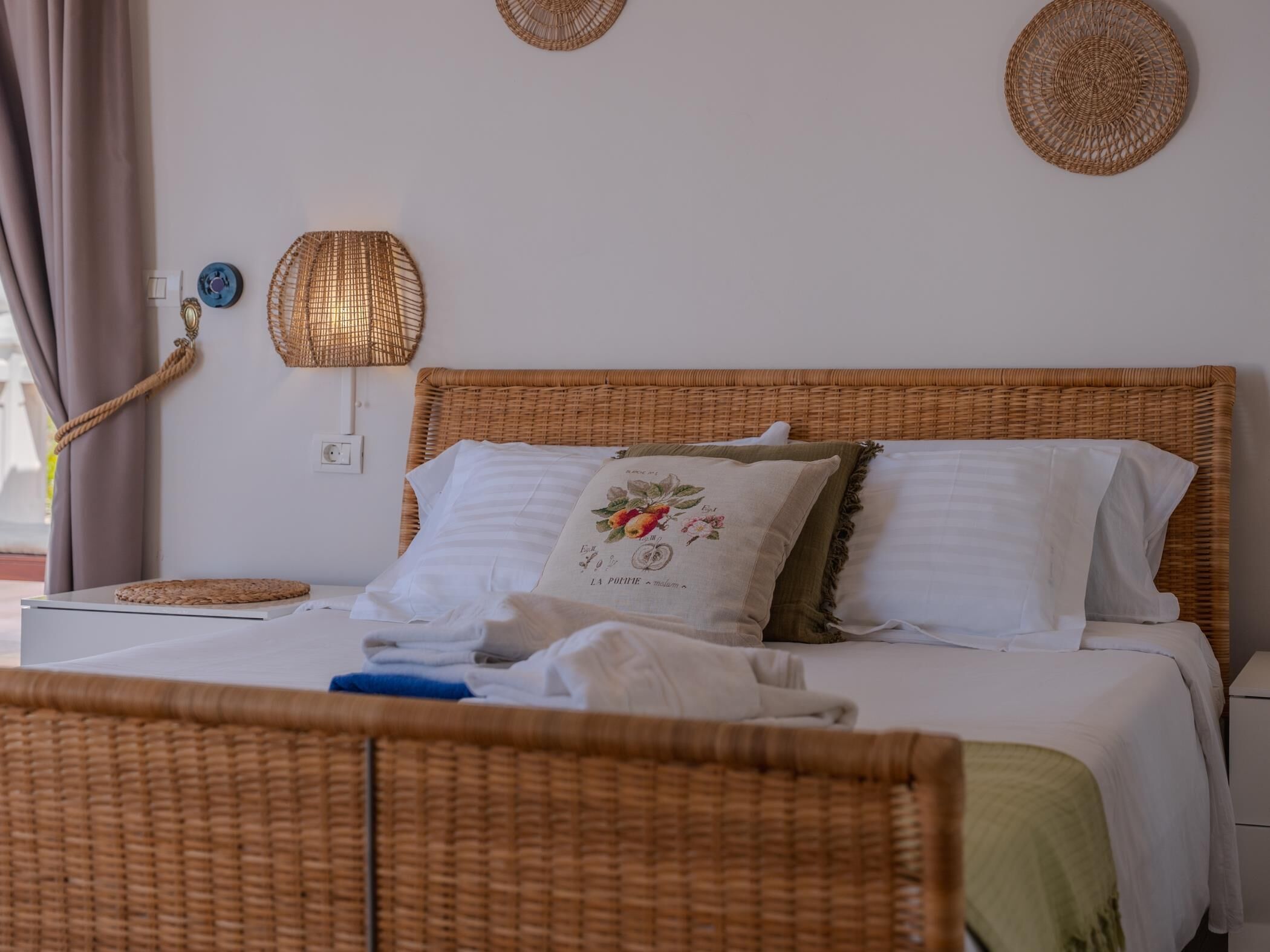 Cama com cabeceira em vime, roupa de cama branca e almofada decorativa com bordado floral.
