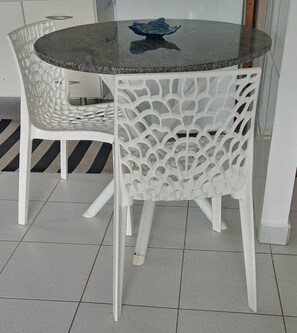 A round table with a dark surface and a blue bowl on top, surrounded by white chairs with a lattice back design. - Ap Beira-Mar - Praia Grande-SP (Praia Grande)