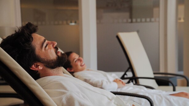 A person lying in a hospital bed with a white blanket, another person sitting beside them, and a chair in the background. - Das Hopfgarten Aparthotel (Hopfgarten im Brixental)