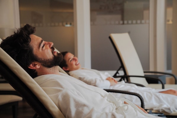A person lying in a hospital bed with a white blanket, another person sitting beside them, and a chair in the background. - Das Hopfgarten Aparthotel (Hopfgarten im Brixental)
