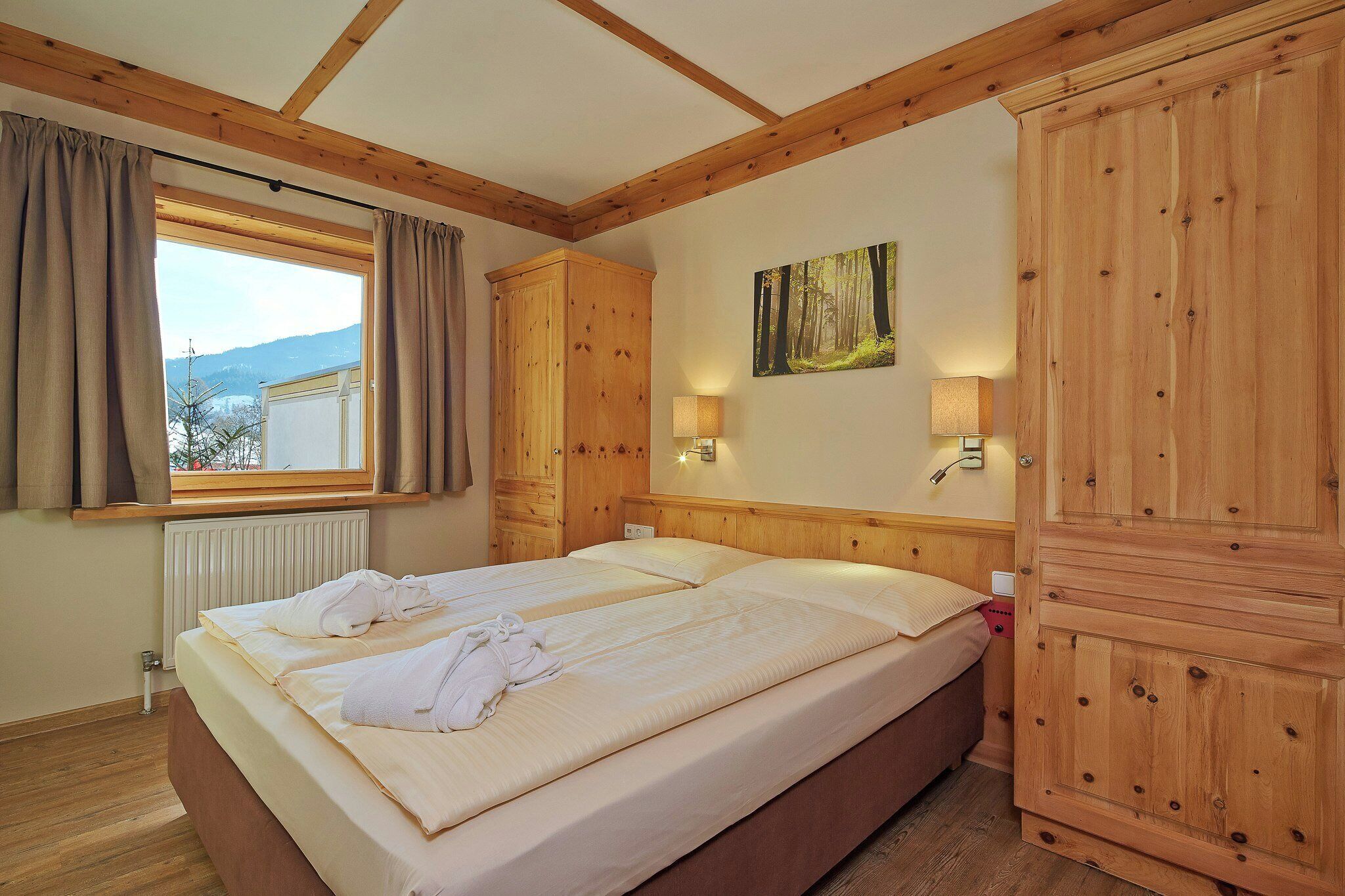 A hotel room with a large bed, wooden headboard, two bedside lamps, a window with curtains, and a painting on the wall.